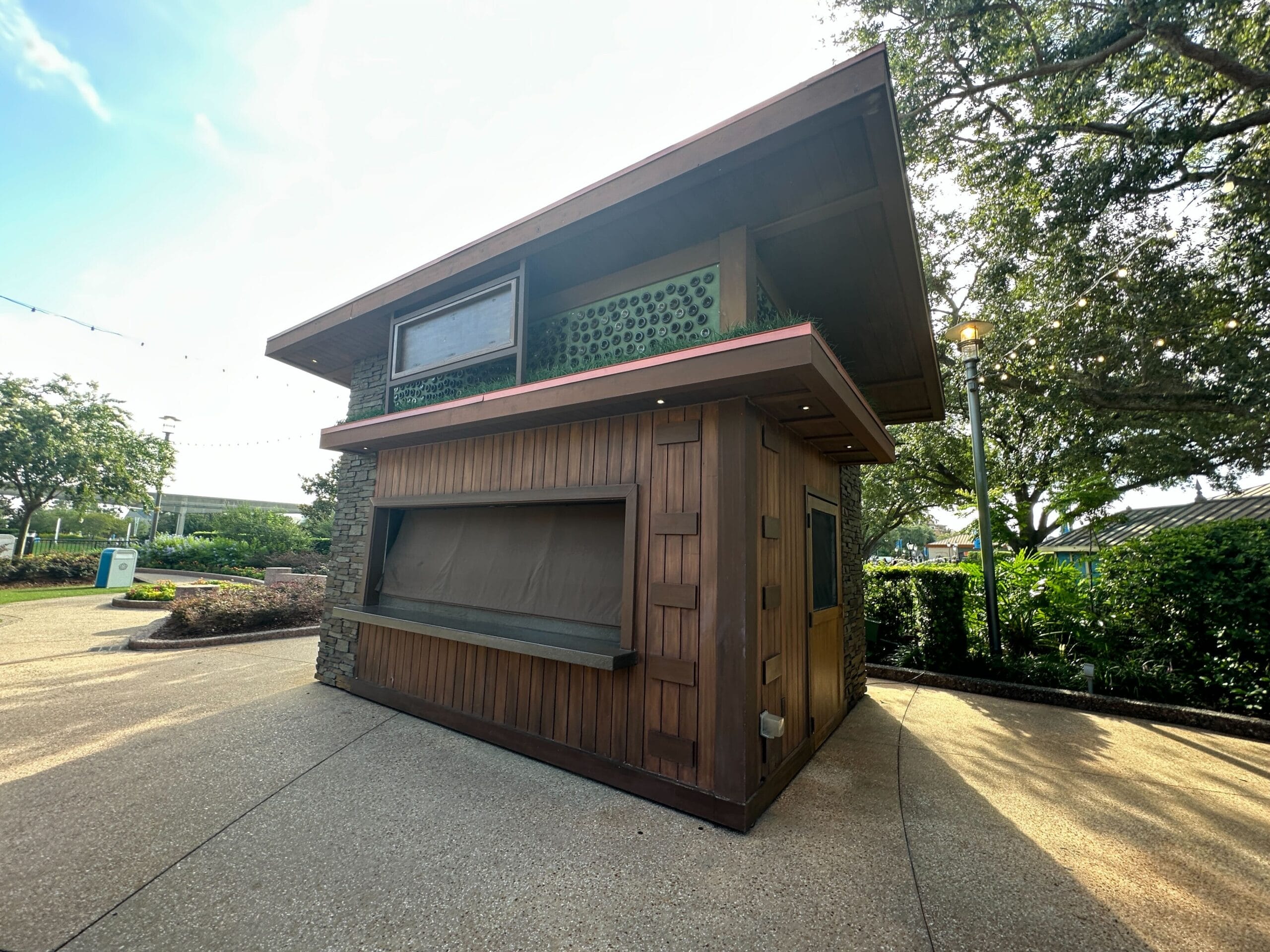 A closed wooden kiosk with shuttered windows stands on a paved path, surrounded by trees and greenery.