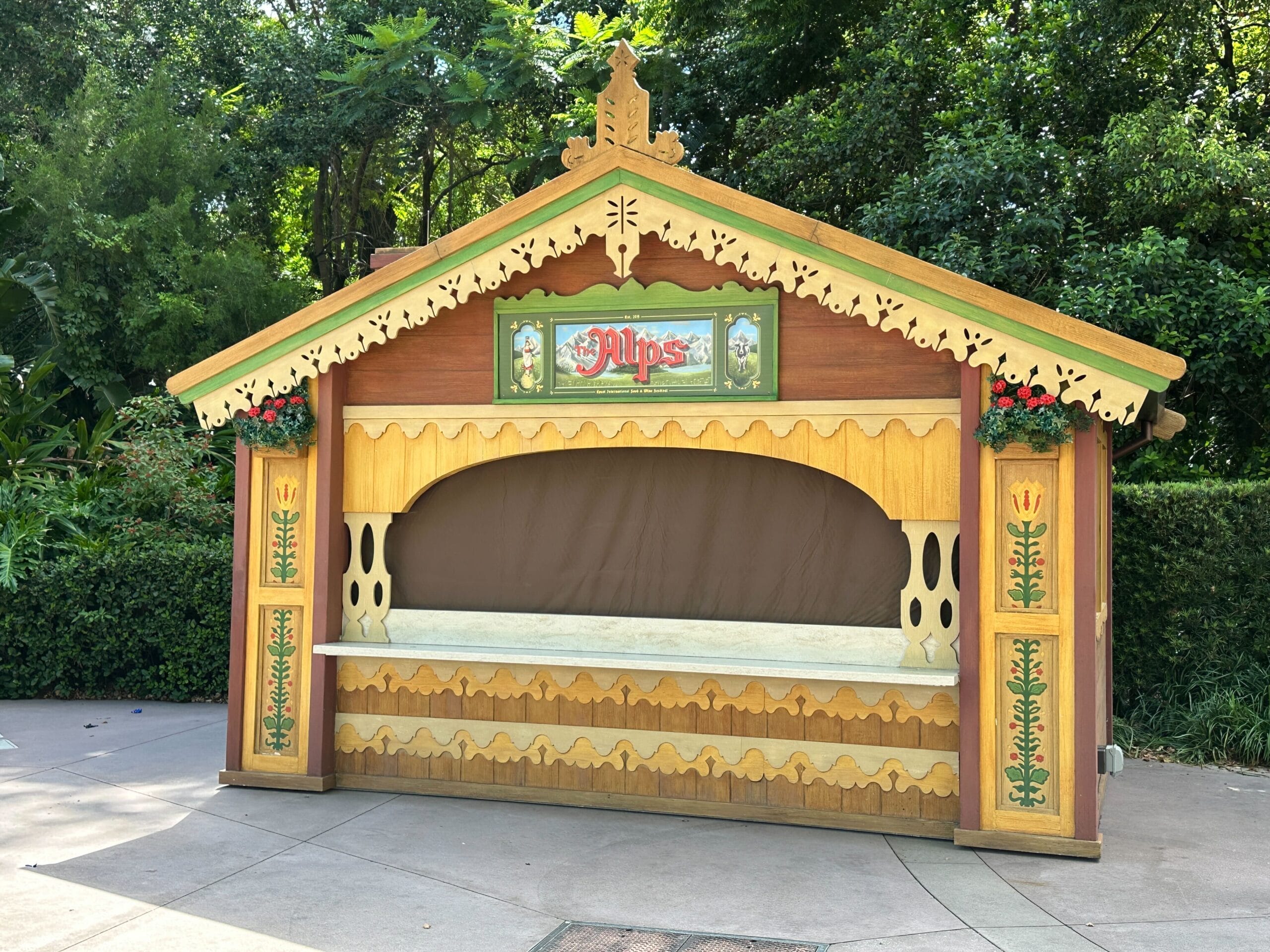 A wooden, chalet-style “Alps” booth stands closed amid greenery, in preparation for the 2025 EPCOT International Food & Wine Festival.